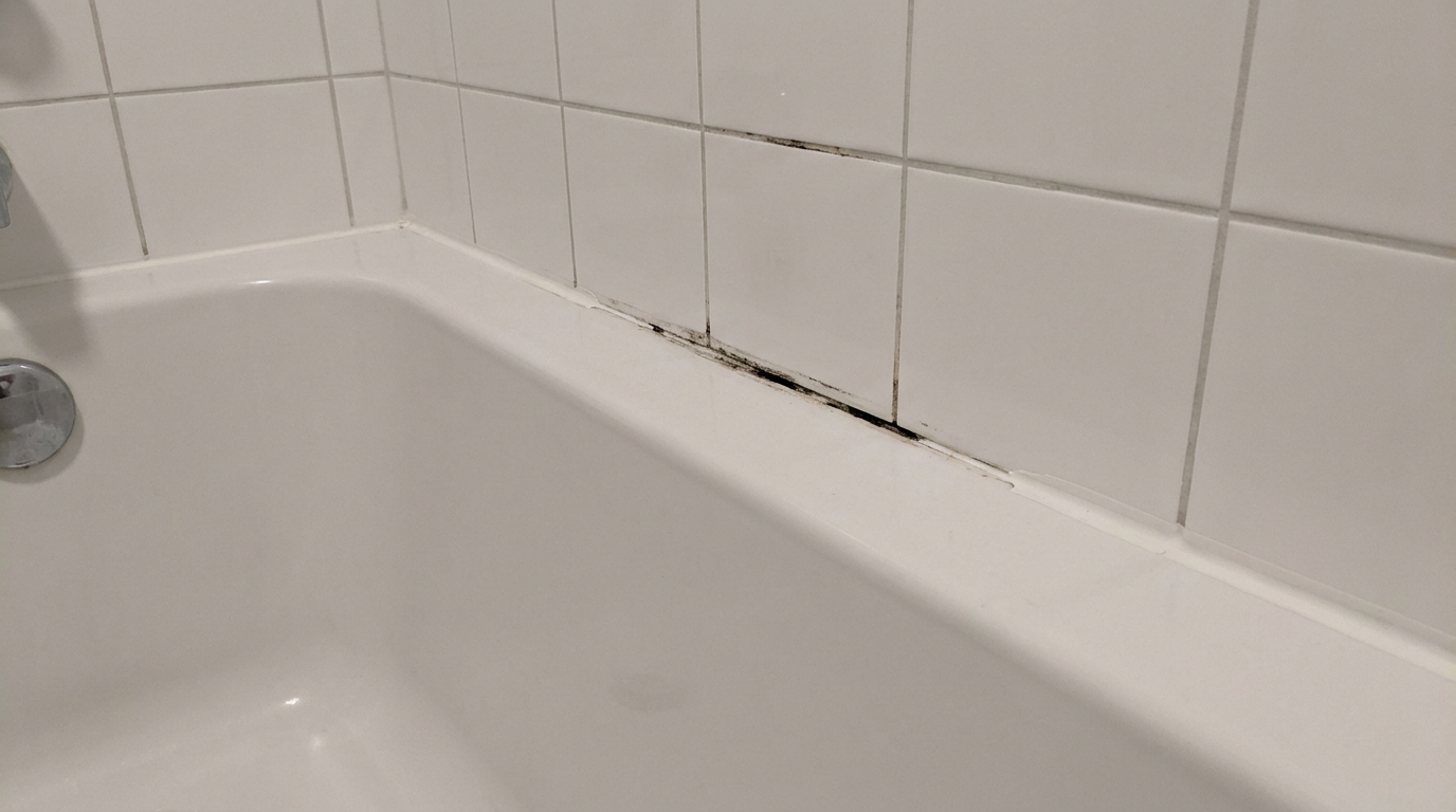 Close-up along the back edge of a white bathtub, where a section of caulk has pulled away from the tile and black mold shows in the gap and along the grout line above it, with the overflow cover at one end of the tub.